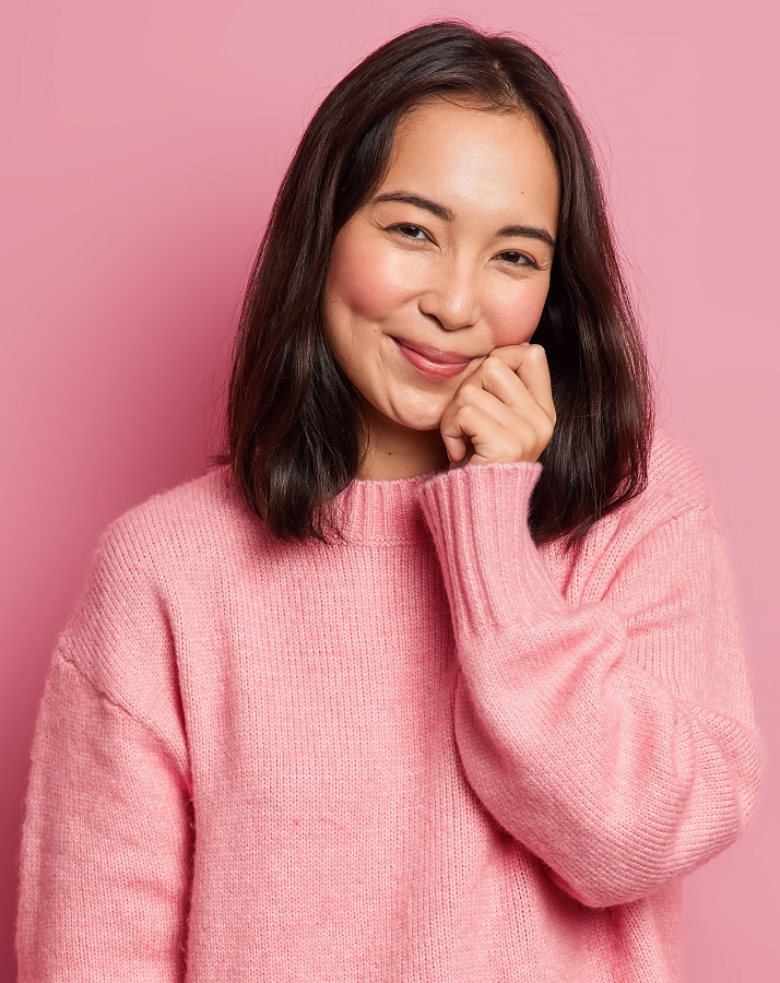 Lovely brunette woman with eastern appearance keeps hand on face has gentle smile looks satisfied at camera has dimples on rouge cheeks poses against pink background. Cute glad Asian girl indoor